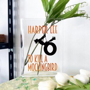 May include: Clear rectangular glass vase with the text "HARPER LEE" and "TO KILL A MOCKINGBIRD" in orange. A black silhouette of a person swinging on a tire swing is in the center. The vase contains green stems and white flowers.