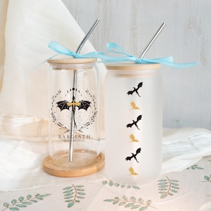 Puede incluir: Dos vasos de vidrio con tapas de bambú y pajitas de metal. Uno es transparente con un diseño negro y dorado y el texto "BASGIATH WAR COLLEGE". El otro es esmerilado con siluetas de dragones negros y dorados. Ambos tienen cintas azul claro.