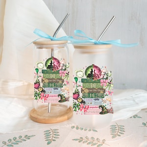May include: Two glass tumblers with bamboo lids and stainless steel straws. The tumblers feature a whimsical design with stacked books, clocks, and a witch silhouette. The design includes the words "Defying Gravity" and "Popular". Each tumbler is tied with a light blue ribbon.