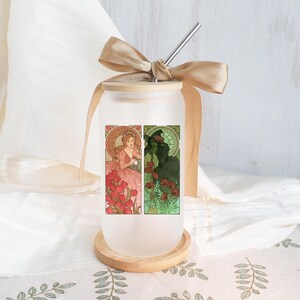 May include: Frosted glass tumbler with a wooden lid, stainless steel straw, and a tan ribbon. The tumbler features two Art Nouveau-style illustrations: one of a woman in a pink dress and the other of a green witch. The tumbler sits on a wooden coaster.