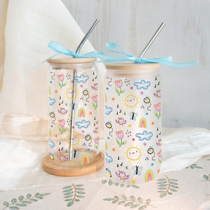 Puede incluir: Dos vasos de cristal con tapas de bambú y lazos azul claro, cada uno con un patrón colorido de soles sonrientes, tulipanes, arcoíris, nubes y abejas. Cada vaso incluye una pajita de metal plateado y se apoya sobre un posavasos de madera.