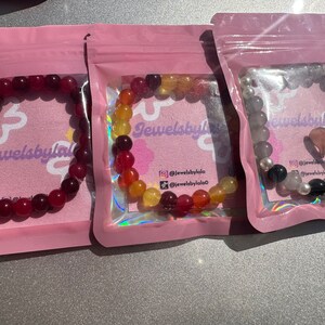 May include: Three bracelets made of colorful beads in pink, red, orange, yellow, and gray. The bracelets are packaged in individual pink pouches with the text "Jewelsbylala" printed on them.