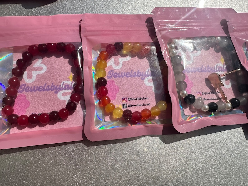May include: Three bracelets made of colorful beads in pink, red, orange, yellow, and gray. The bracelets are packaged in individual pink pouches with the text "Jewelsbylala" printed on them.