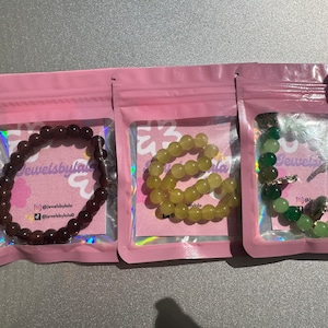 May include: Three bracelets made of different colored beads in individual pink packaging. The bracelets are labeled "Jewelsbylala" and have a social media handle.