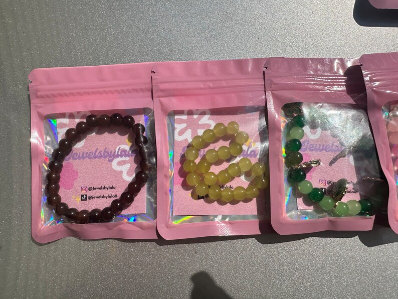 May include: Three bracelets made of different colored beads in individual pink packaging. The bracelets are labeled "Jewelsbylala" and have a social media handle.