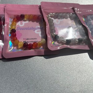 May include: Four small bags with pink and clear plastic packaging containing colorful beaded bracelets. The bracelets are made of red, orange, yellow, gray, and purple beads. The bags have the text "Jewelsbylala" printed on them.