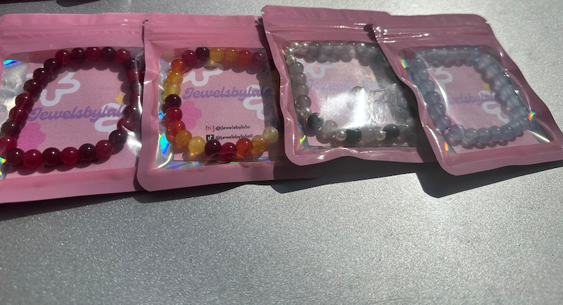 May include: Four small bags with pink and clear plastic packaging containing colorful beaded bracelets. The bracelets are made of red, orange, yellow, gray, and purple beads. The bags have the text "Jewelsbylala" printed on them.
