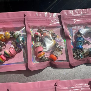 May include: Three pink zippered pouches with clear plastic windows, each containing a colorful charm bracelet. The bracelets feature a variety of beads, including hearts, flowers, and animals.