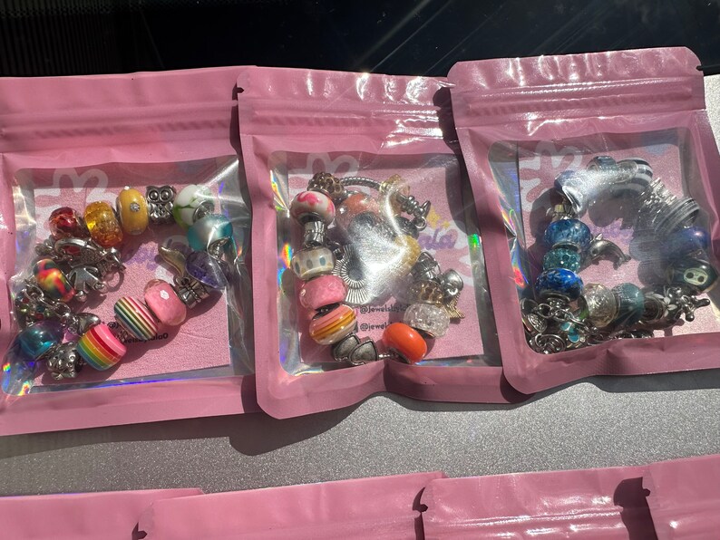 May include: Three pink zippered pouches with clear plastic windows, each containing a colorful charm bracelet. The bracelets feature a variety of beads, including hearts, flowers, and animals.