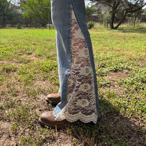 Puede incluir: Vaqueros de mezclilla azul claro con un panel de encaje color crema en el lateral. Los vaqueros se llevan con botas de cuero marrones. El encaje añade un elemento decorativo al estilo denim clásico.