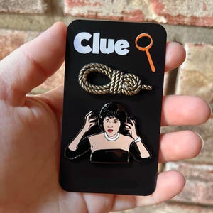 May include: A black card with the word "Clue" in white letters and a magnifying glass icon. The card features three enamel pins: a gold rope, a woman with black hair and a black dress, and a magnifying glass.