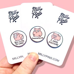 May include: Three round enamel pins with white borders and pink hamster illustrations. One pin reads "Too Cute To Care", another says "Social battery: 0%", and the third says "I Hate Everyone". The pins are on white cards with the "Milly Pins" logo.
