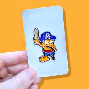 May include: A cartoon orange cat wearing a blue pirate hat with a skull and crossbones design, holding a wooden sword. The cat is wearing a red sash and blue shorts. The pin is on a white background.