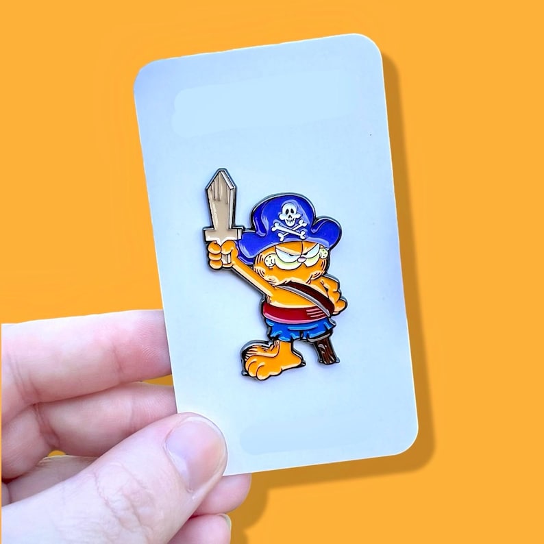 May include: A cartoon orange cat wearing a blue pirate hat with a skull and crossbones design, holding a wooden sword. The cat is wearing a red sash and blue shorts. The pin is on a white background.