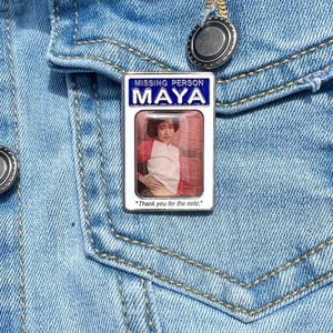 May include: A rectangular enamel pin with the text "MISSING PERSON MAYA" in blue and white. The pin features a photo of a person in a red and white shirt. The text "Thank you for the note." is at the bottom. The pin is attached to a denim jacket.