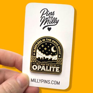 May include: A black and gold enamel pin with the text "Sleepless in the Onyx Night" and "But Now The Sky Is Opalite." The pin features a night sky design with stars and a crescent moon. The pin is on a white card with the text "Pins by Milly."