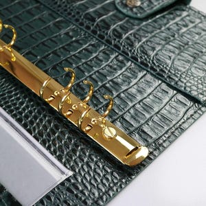 K&ouml;nnte beinhalten: Ein dunkelgr&uuml;ner Planer-Einband mit texturiertem Krokodilleder-Muster. Das Innere verf&uuml;gt &uuml;ber eine goldfarbene Sechs-Ring-Bindemechanik aus Metall und ein transparentes Kunststoff-Kartenfach auf der linken Seite. Der Einband hat einen Druckknopfverschluss.