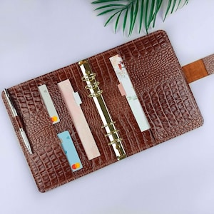 May include: A brown leather planner with a crocodile pattern, gold rings, and a brown leather flap closure. The planner is open and has several cards and a piece of paper inside.