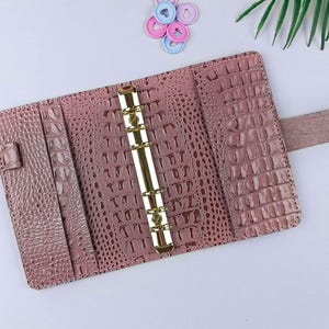 May include: A dusty rose colored personal planner binder featuring a textured crocodile skin pattern. The interior includes a gold-toned six-ring metal mechanism, vertical storage pockets on the left side, and a matching strap with a snap closure.