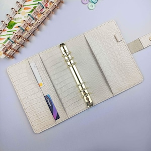 May include: A white faux leather planner with a gold ring binder and a crocodile embossed pattern. The planner has a card slot and is open to reveal the gold rings.