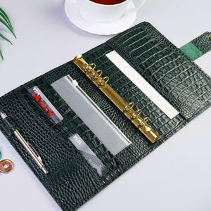 K&ouml;nnte beinhalten: Ein dunkelgr&uuml;ner Planer-Einband mit Krokodilleder-Pr&auml;gung, ausgestattet mit einer goldfarbenen Sechs-Ring-Mechanik, einer Stiftschlaufe mit silbernem Stift, mehreren Kartenf&auml;chern mit Visa-Karten, einem transparenten Einsteckfach und einem Verschlussriemen mit Druckknopf.