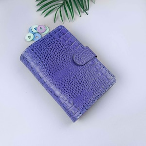 May include: A purple faux-leather planner with a crocodile skin texture. The planner is closed with a matching snap closure. Small, colorful disc-shaped planner accessories are visible in the background.