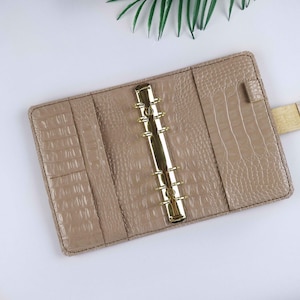 May include: A beige leather bound planner with a gold metal ring binder. The planner has a crocodile embossed pattern.
