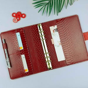 May include: Open red faux-leather planner with a snakeskin texture. The planner has a gold binder, a pen, and several inserts. A red strap with a snap closure secures the planner. Small red circular objects are in the upper left corner.