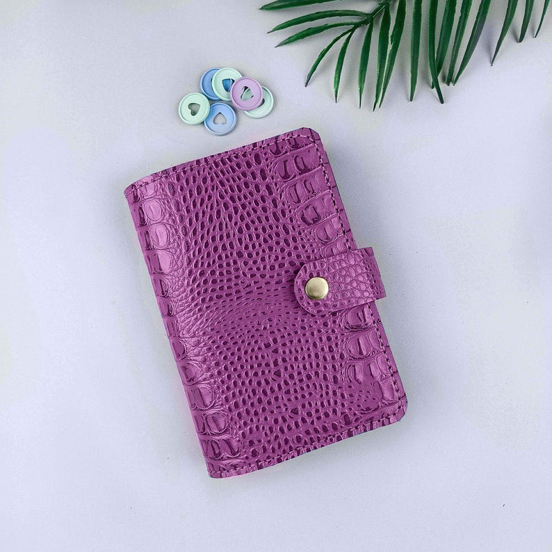 K&ouml;nnte beinhalten: Ein violettes Notizbuch mit einer strukturierten Krokodilleder-Optik und einem goldfarbenen Druckknopfverschluss. Das Notizbuch wird von einer kleinen Gruppe aus f&uuml;nf pastellfarbenen Kunststoffscheiben begleitet, die mittig herzf&ouml;rmig ausgestanzt sind.
