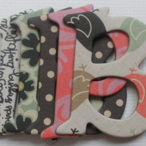 May include: A set of fabric letters in the shape of the letter "B" with a patchwork design. The letters are made of different fabrics with various patterns, including floral, polka dot, and geometric designs. The letters are in shades of pink, green, black, and white.