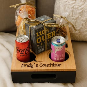 Könnte beinhalten: Ein Couch-Tablett aus Bambus mit zwei Getränkehaltern, einem Griff und dem Text "Andy's Couchbar". Es enthält eine Coca-Cola-Dose, eine rosa Dose, eine Schachtel und zwei Snack-Tüten. Auf der Schachtel steht "110% QUER DR."