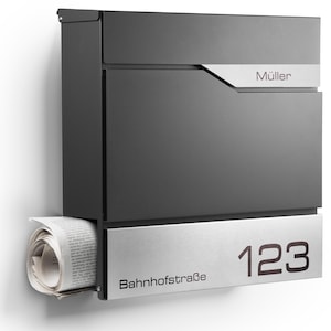 Puede incluir: Un buzón moderno de color gris oscuro con un acento plateado y el nombre "Müller" impreso. El buzón tiene un compartimento para periódicos, con un periódico enrollado visible. El número "123" y el nombre de la calle "Bahnhofstraße" se muestran.