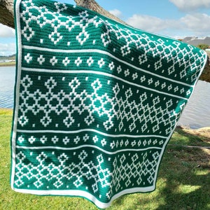 May include: A handmade, green and white crochet blanket with a geometric pattern. The blanket is draped over a tree branch, showcasing its intricate design and texture. The white border frames the detailed pattern.