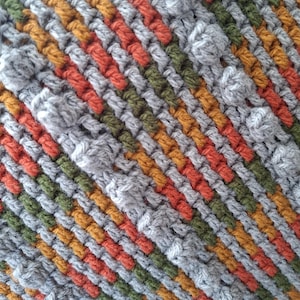 Peut inclure: Un textile crocheté présentant un motif diagonal répétitif de fils dans des tons de gris, vert olive, jaune moutarde et orange brûlé. Le tissu affiche une surface texturée avec des points popcorn intégrés dans les rangées de fil gris.