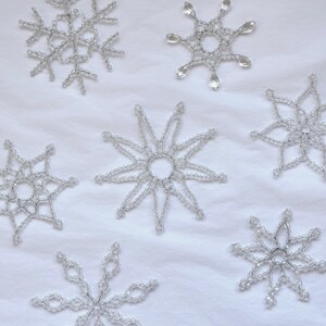 Peut inclure: Sept flocons de neige perlés transparents de différents modèles sont disposés sur une surface blanche. Les flocons de neige sont faits de perles transparentes assemblées pour former des formes symétriques complexes, parfaites pour la décoration hivernale.