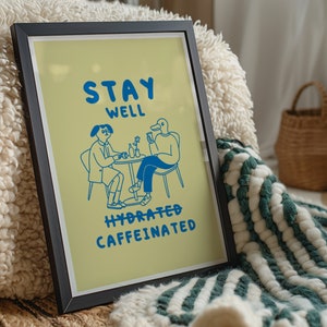 Puede incluir: Una impresión enmarcada con un fondo amarillo e ilustraciones azules de dos personas sentadas en una mesa. El texto en la impresión dice "Stay Well Hydrated Caffeinated".