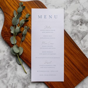 May include: A wedding menu card with a light blue font on a white background, listing starters, main courses, and dessert options. The menu is on a wooden cutting board with eucalyptus leaves.