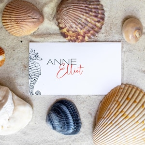May include: A white business card with black text that reads "Anne Elliot" and a black and white illustration of a seahorse. The card is surrounded by seashells on a sandy background.