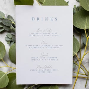 May include: A white menu card with the word "DRINKS" in a blue font. The card lists drink options: Beer & Cider, Wine, Spirits, and Non-Alcoholic beverages. Eucalyptus leaves frame the card.