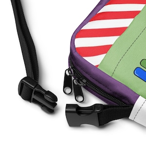 May include: A close-up of a colorful fanny pack with a black strap and buckle. The bag features a red and white striped pattern, a green section, and a purple zipper. The black zipper pulls are visible.