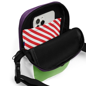 May include: A small, open crossbody bag with a purple top, white, green, and black accents. The bag has a black strap and zipper. Inside, a phone and a red and white striped item are visible.