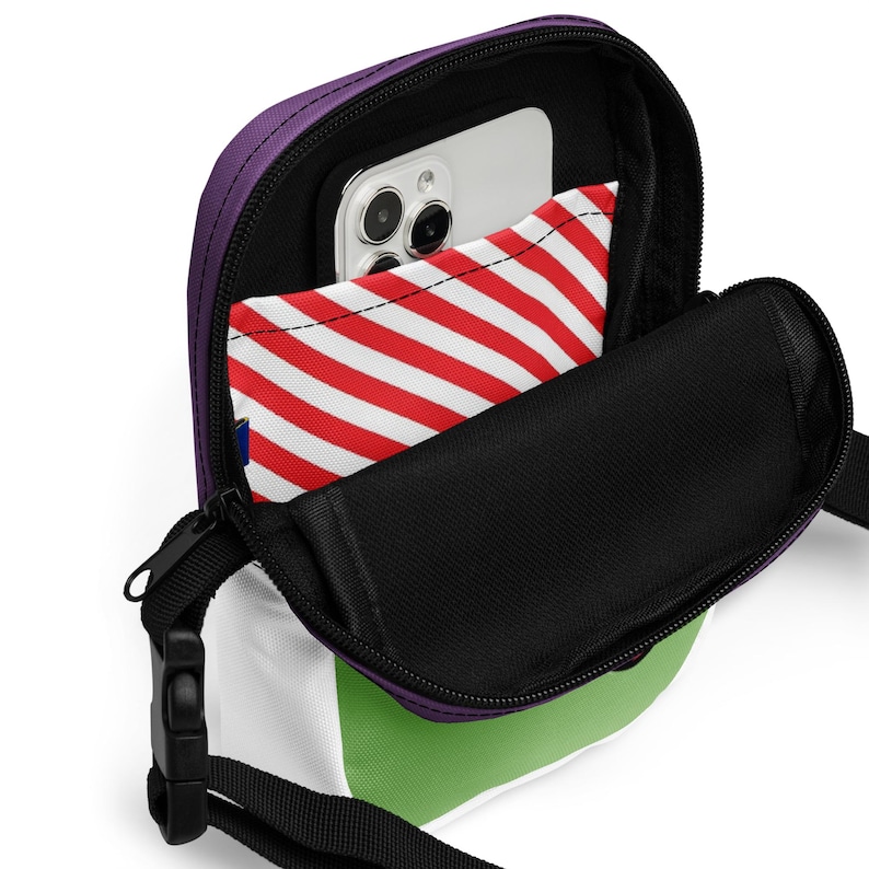 May include: A small, open crossbody bag with a purple top, white, green, and black accents. The bag has a black strap and zipper. Inside, a phone and a red and white striped item are visible.