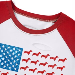 May include: White and red raglan-style t-shirt with a design combining the American flag with red dachshunds. The flag's blue section has glittery stars. The shirt has a distressed look with a small tear.