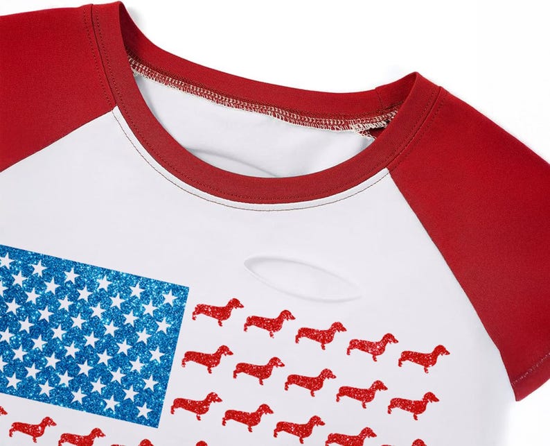 May include: White and red raglan-style t-shirt with a design combining the American flag with red dachshunds. The flag's blue section has glittery stars. The shirt has a distressed look with a small tear.