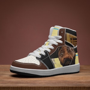 May include: A high-top sneaker featuring a brown and white color scheme with black accents. The side panel displays a graphic illustration of a person with wide eyes and a beard. The shoe includes white laces, a perforated toe box, and a white sole.