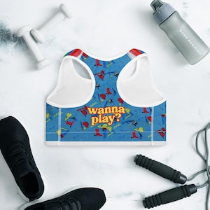 Op de afbeelding: Witte sportbeha met een blauw-wit patroon. Het patroon bevat verschillende kleurrijke cartoonafbeeldingen van gereedschap en de tekst "wanna play?" in gele en oranje letters.