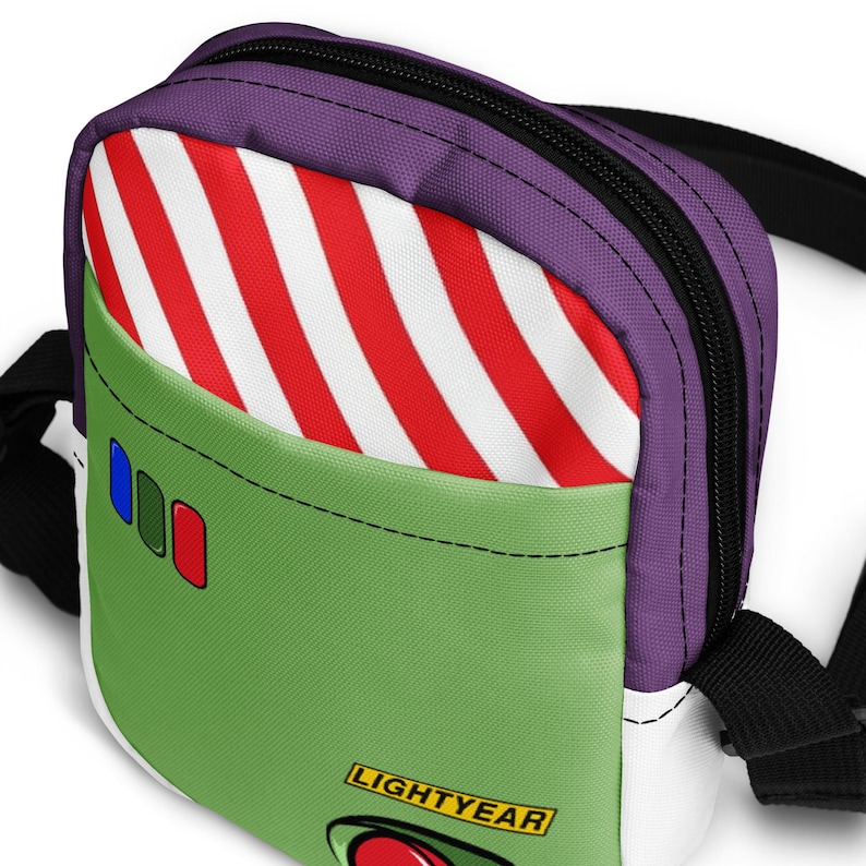 May include: A small, rectangular crossbody bag with a design inspired by the character Buzz Lightyear. The bag features a green front panel, a red and white striped pocket, and a purple top. The text "LIGHTYEAR" is visible on the front.