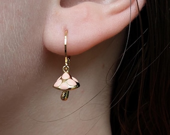 May include: A gold-colored earring with a mushroom charm. The mushroom cap is light pink with white spots, and the stem is gold. The earring is attached to a gold hoop that is inserted into the ear.