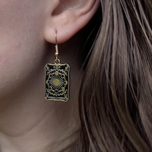 May include: A gold-colored earring with a black rectangular pendant. The pendant features a gold sun and moon design with the text "THE WHEEL OF FORTUNE". The earring is hanging from a gold-colored hook.