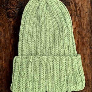 Può includere: Un berretto lavorato a maglia verde chiaro con bordo ripiegato. Il cappello è realizzato in tessuto a maglia a coste e viene mostrato su una superficie di legno scuro. Il cappello è in tinta unita e ha un design semplice e classico.